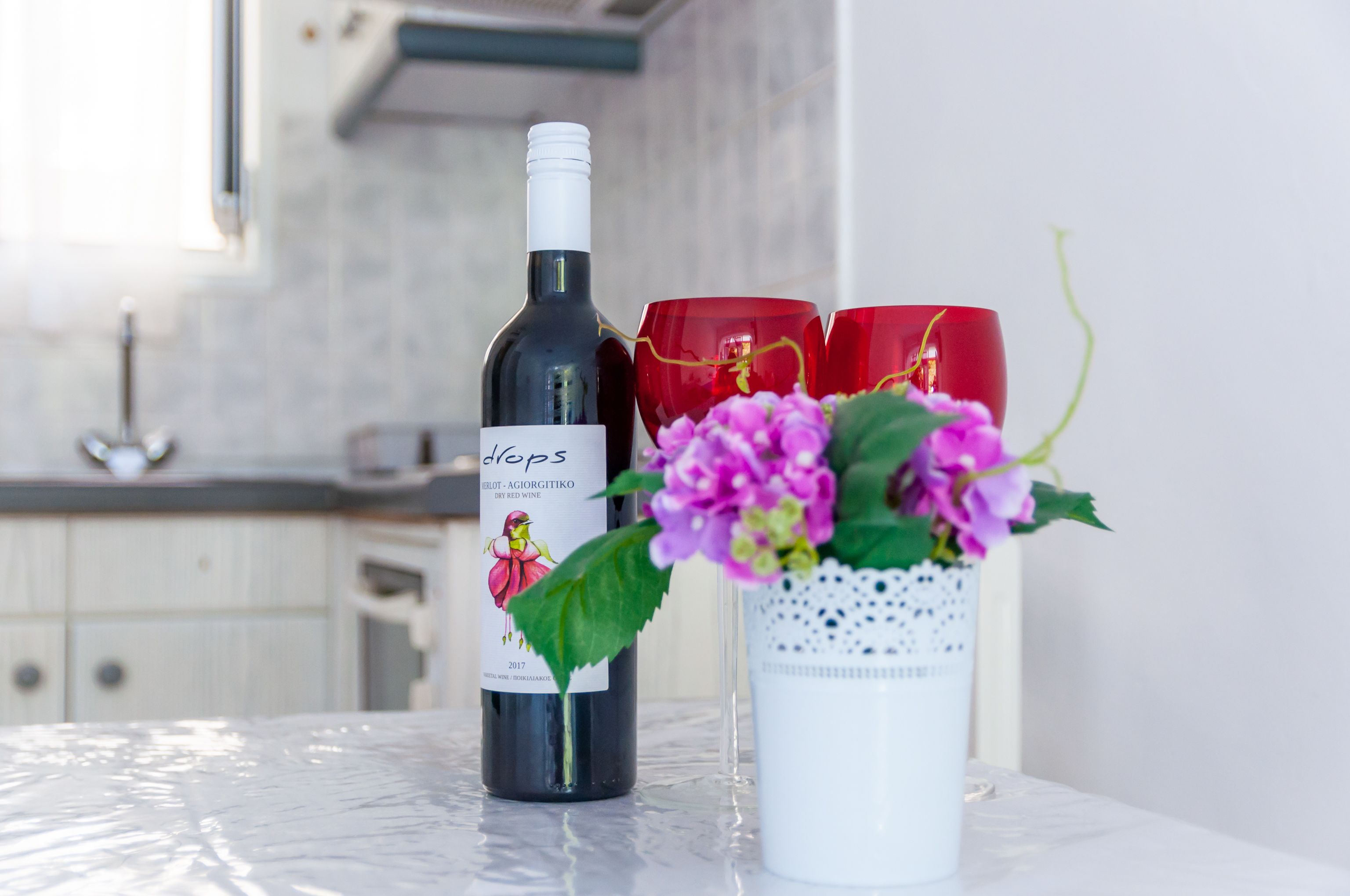 A bottle of wine with a label, two red glasses, and a white vase with purple flowers on a table.