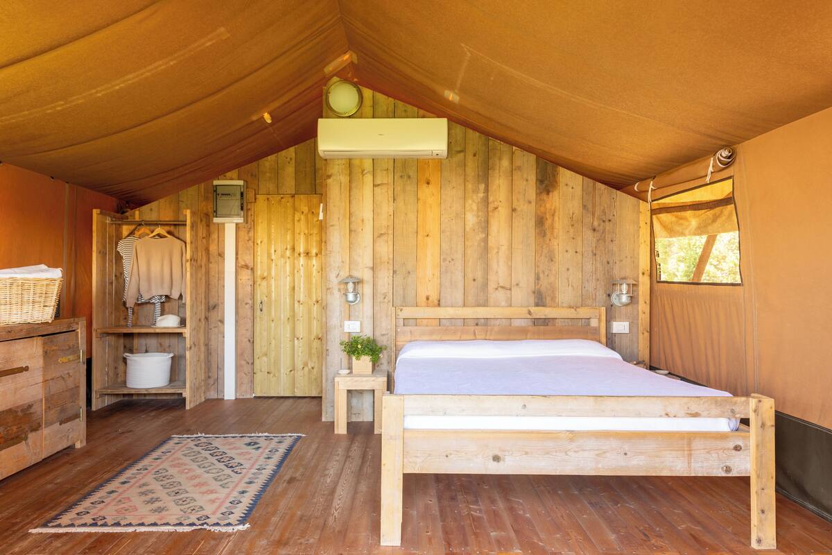 Une pièce en bois aménagée comme une tente, avec un lit, une petite table, un porte-vêtements suspendu et une fenêtre.