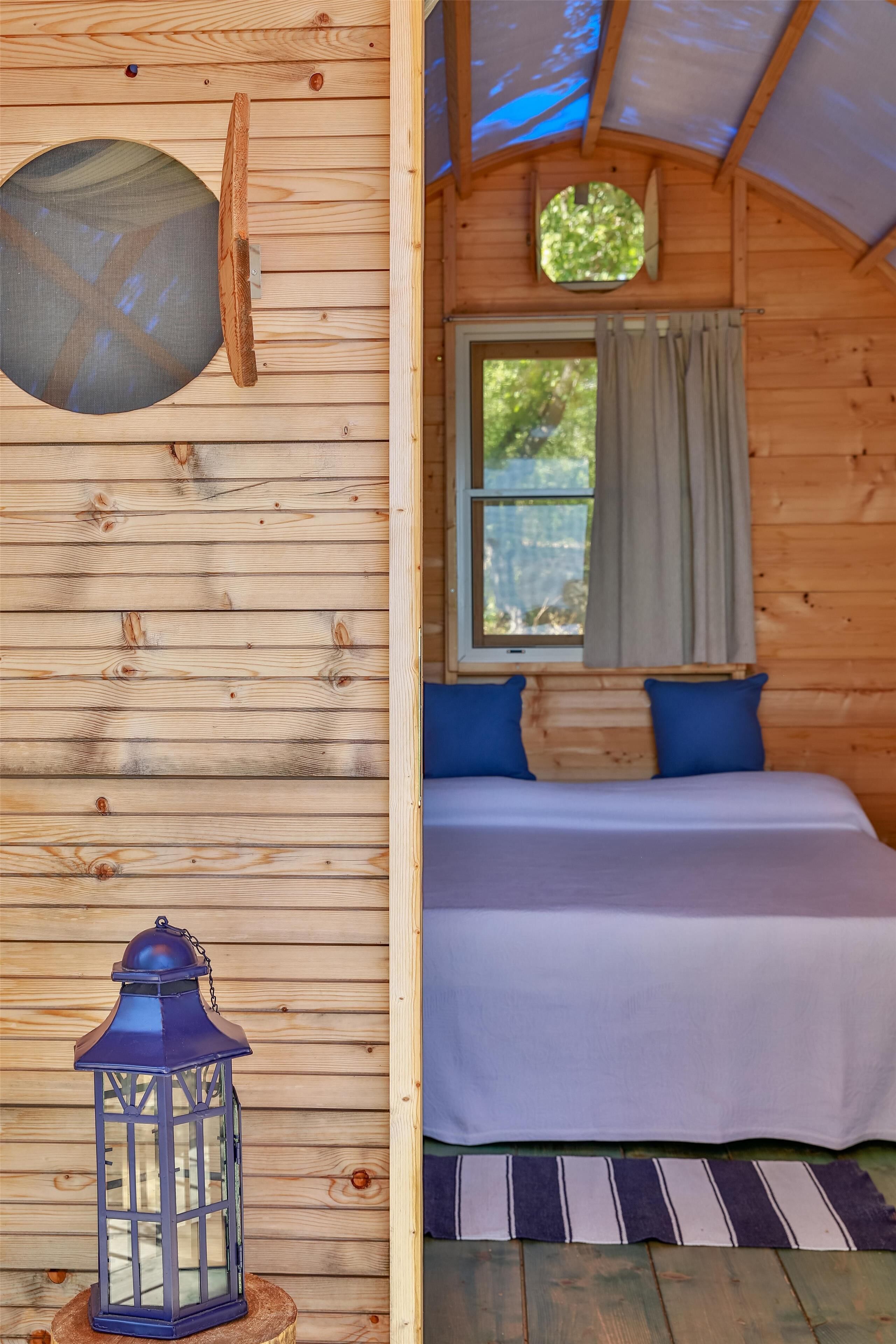 A wooden cabin interior with a bed, a window with curtains, a lantern, and a circular skylight.