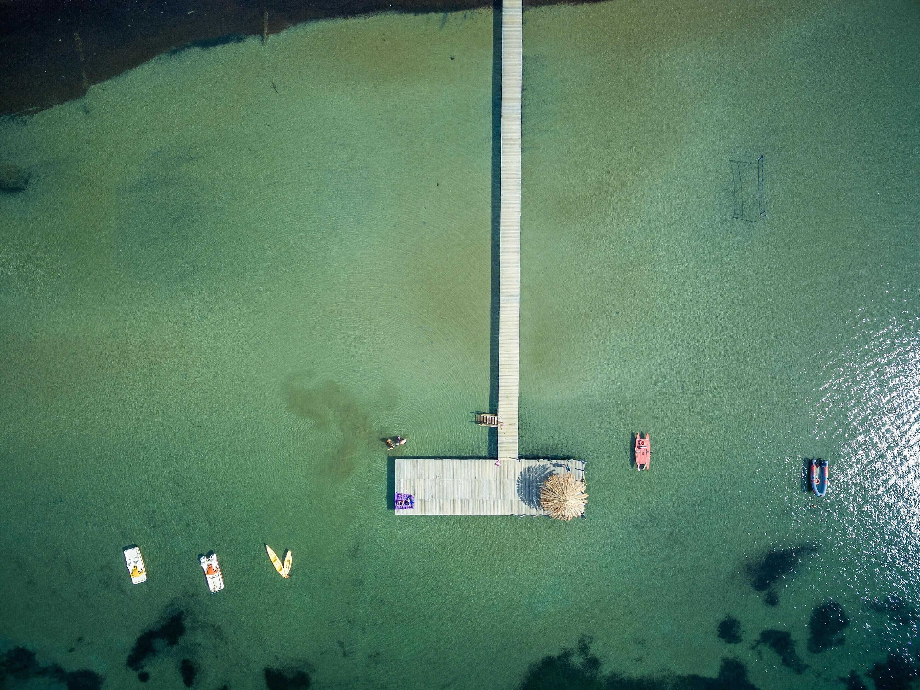 An aerial view of a dock with a pier extending into a body of water. There are several boats and a small structure with a flag on the pier.