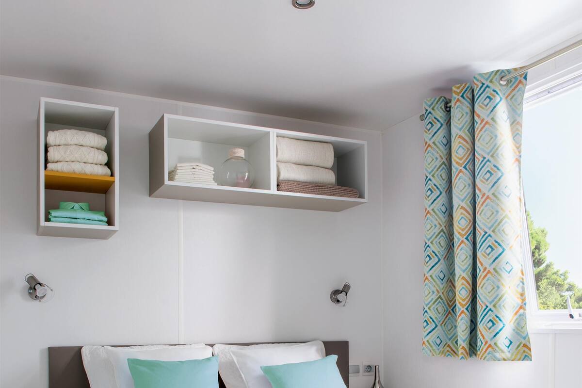 Un lit bien fait, avec un plateau contenant des tasses et une carafe, une table de chevet et une étagère où sont pliées des serviettes.