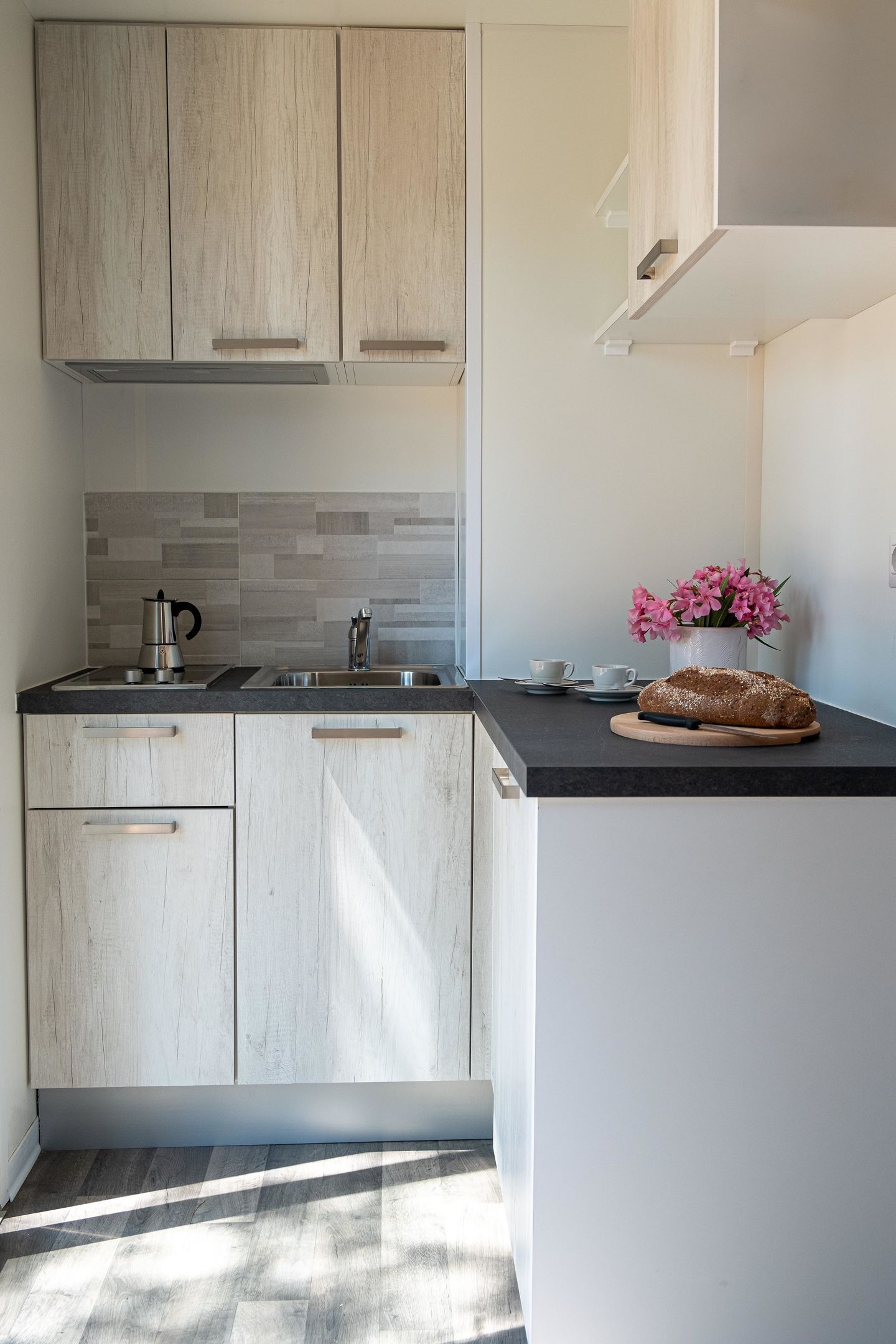 A modern kitchen with light wood cabinets, a black countertop, a stainless steel sink, a kettle, a cup, and a loaf of bread.