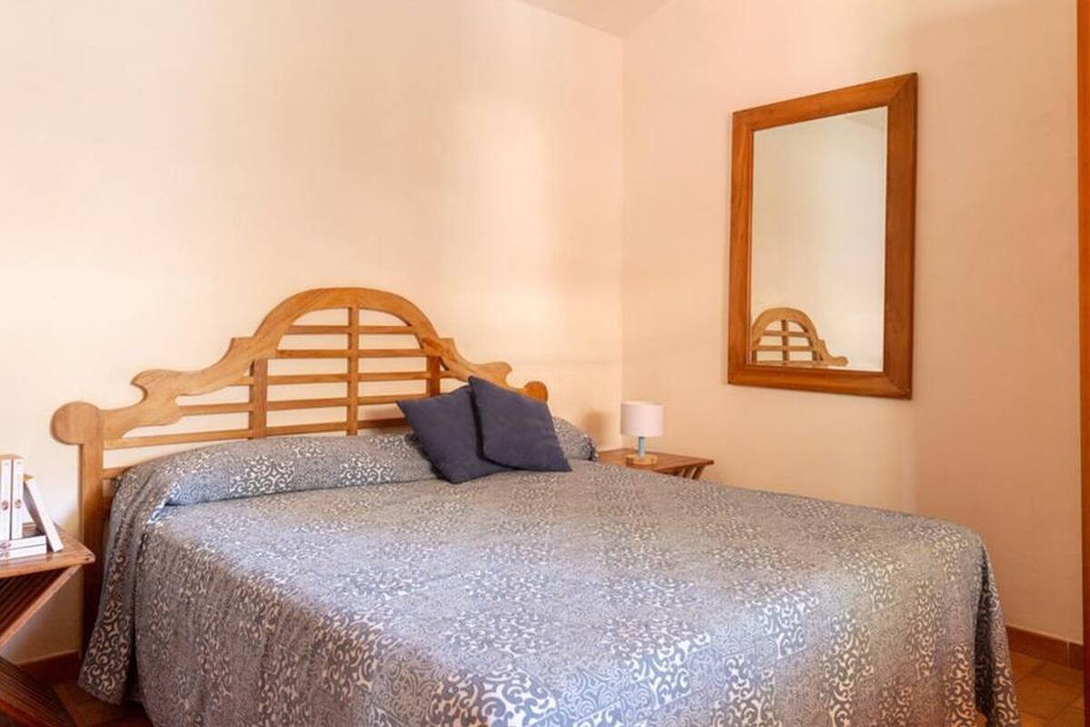 Une chambre avec une tête de lit en bois, une couvre-lit à motifs, un seul oreiller, une table de chevet avec une lampe et un miroir sur le mur.