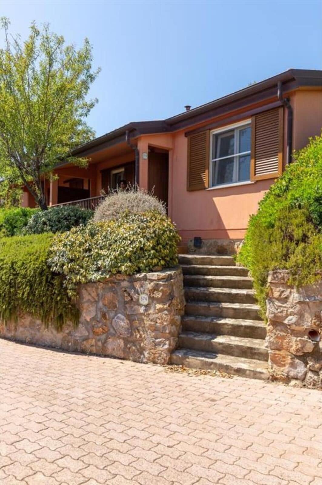 A two-story house with a stone staircase leading to the front door, surrounded by well-maintained hedges and a tree.