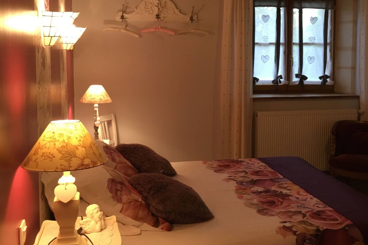 Une chambre à coucher comprenant un lit, deux lampes de chevet, un élément décoratif mural, une fenêtre avec des rideaux et un radiateur.