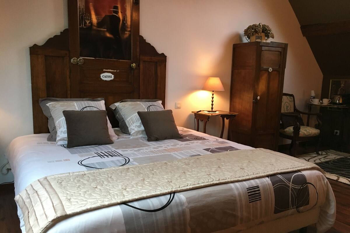 Un lit bien fait, recouvert d’une couvre-lit à motifs et agrémenté d’oreillers, une armoire en bois avec un miroir, une petite table avec une lampe et une chaise, le tout dans une pièce au plancher en bois.
