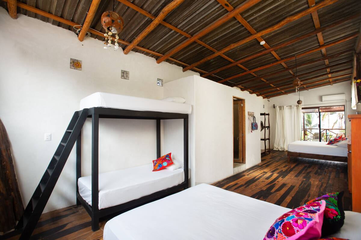 A room with a bunk bed, a single bed, wooden flooring, a rustic ceiling, and a window with curtains.