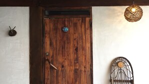 Una porta de fusta amb mànec metàl·lic, una planta en test i una cadira de vimet davant d'un edifici.