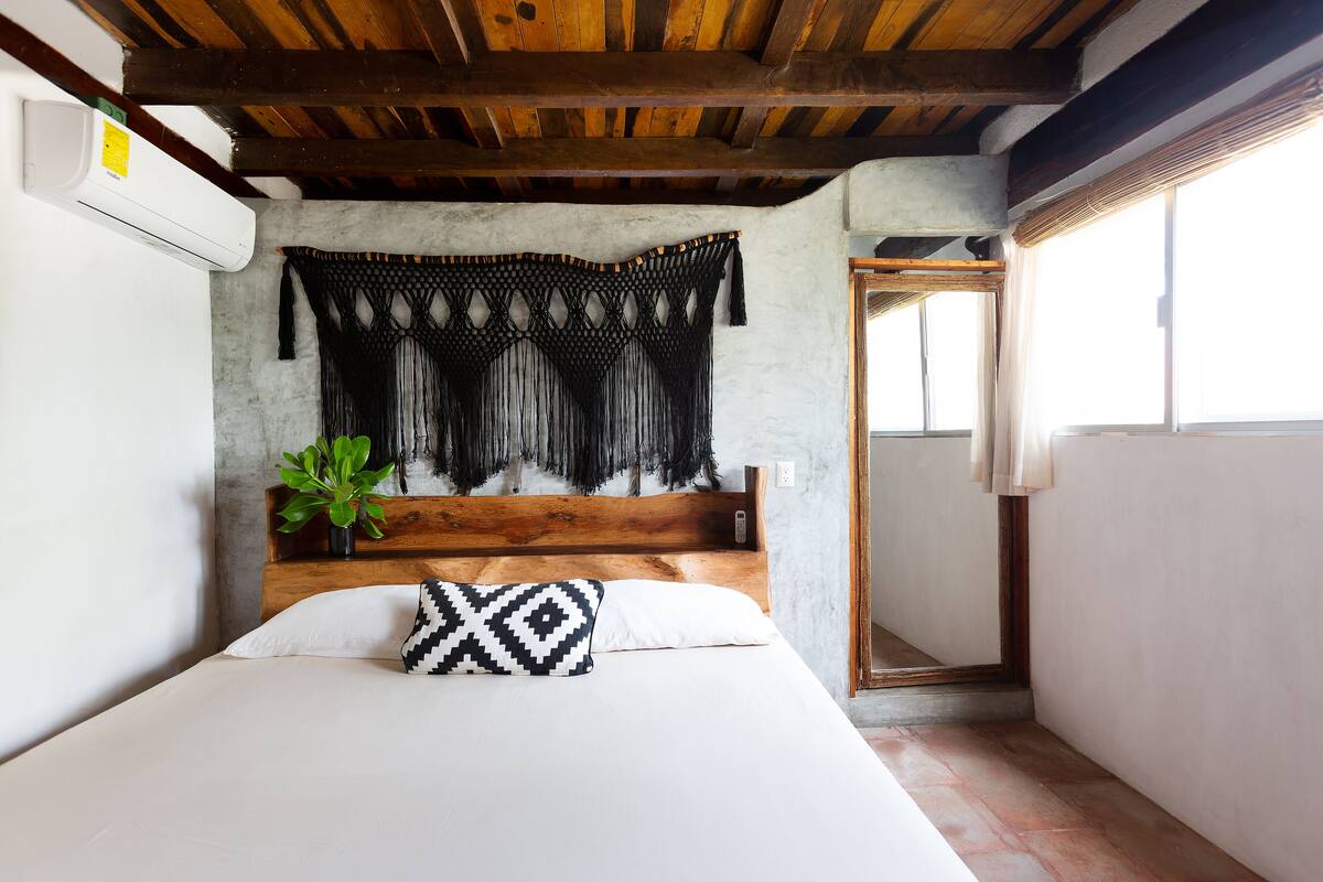 A bedroom with a wooden headboard, a large window, and a macramé wall hanging.