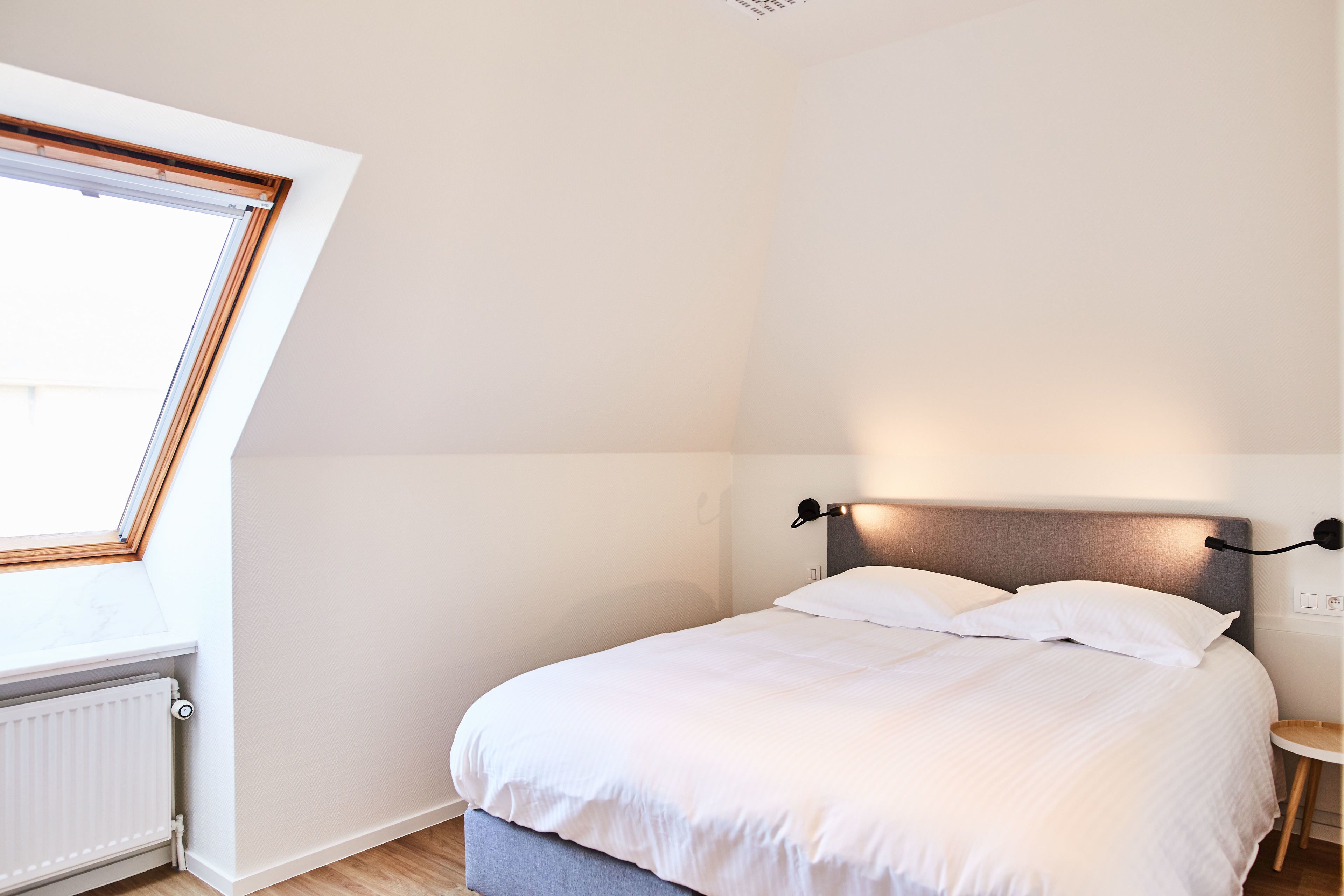A bedroom with a bed, a nightstand, a wall-mounted lamp, a radiator, and a window with a wooden frame.