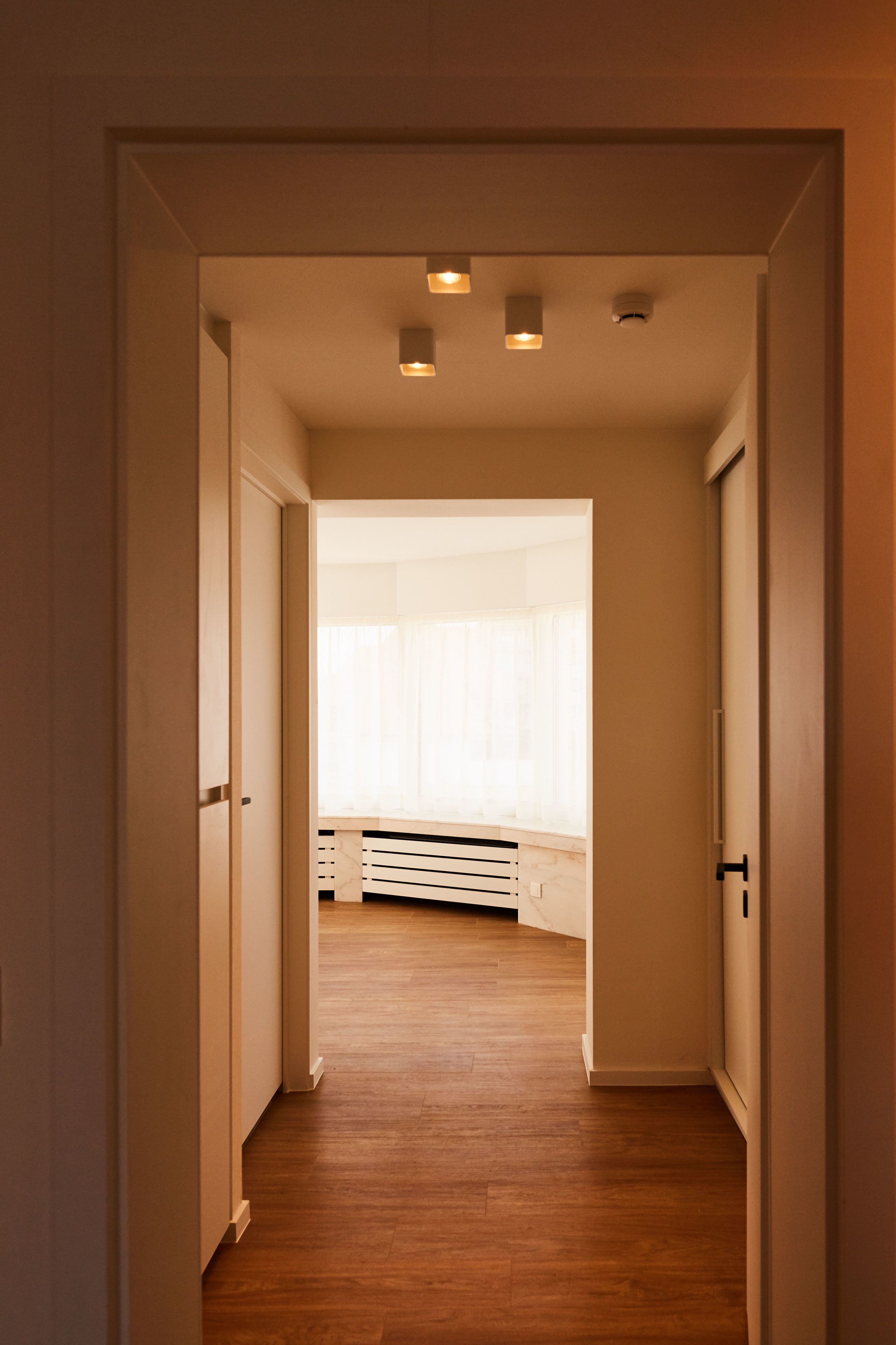 A hallway with wooden flooring leading to a room with a large window and a bed.
