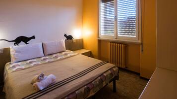 A hotel room with a bed, two pillows, a blanket, a wall decoration of a cat, a window with blinds, and a radiator.