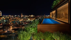 Zona de piscina en la azotea con plantas en macetas y velas, con vistas a un paisaje urbano nocturno.