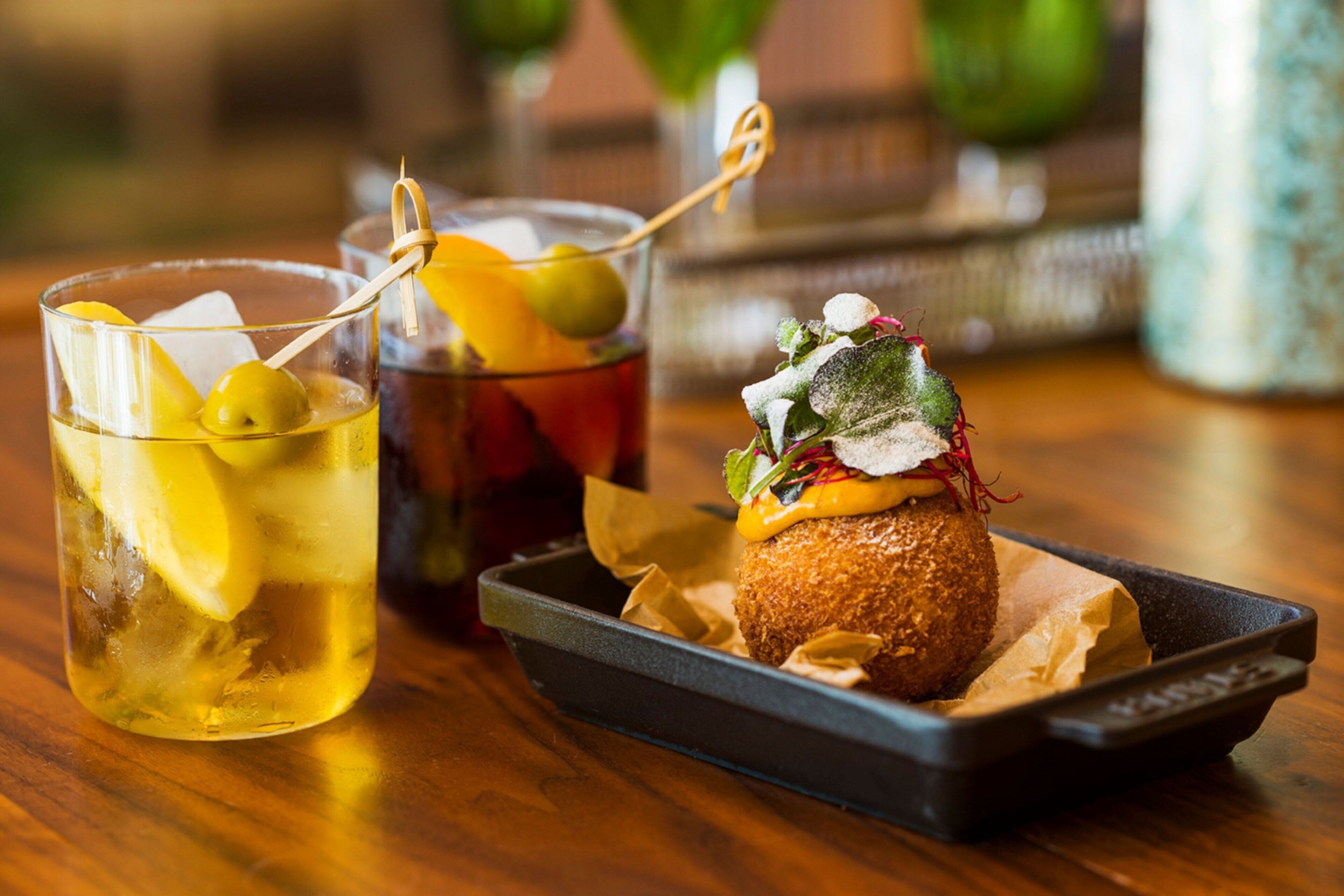 A tray with a breaded item topped with vegetables and cheese, accompanied by two glasses of iced beverages with fruit slices and olives.