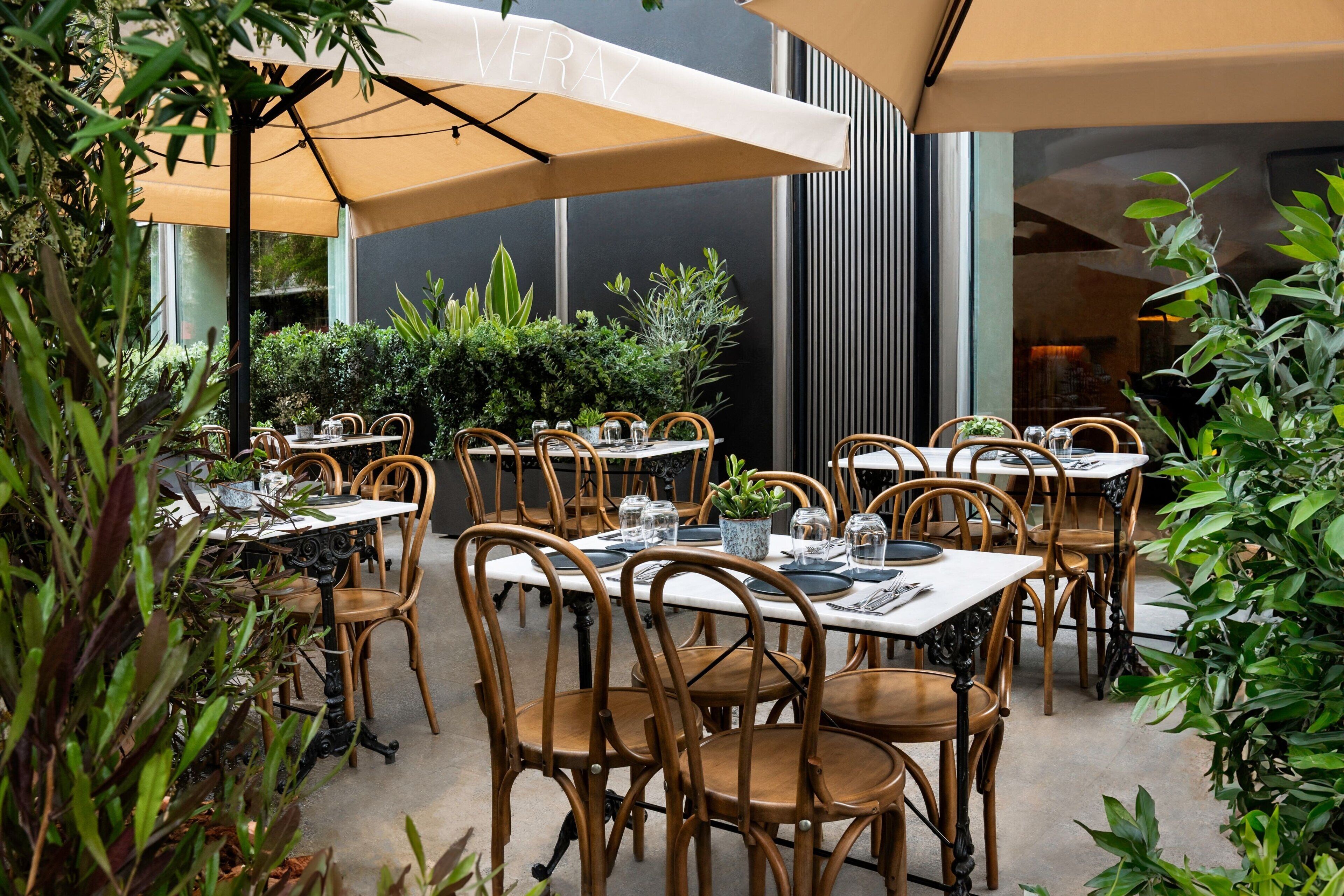 Un área de comedor al aire libre con mesas y sillas, rodeada de follaje y cubierta por sombrillas.