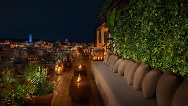 Terraza en la azotea con vista a la ciudad de noche, con una fila de sofás y plantas en macetas.
