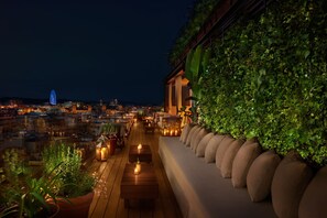 Terraza en la azotea con vista a la ciudad de noche, con una fila de sofás y plantas en macetas.