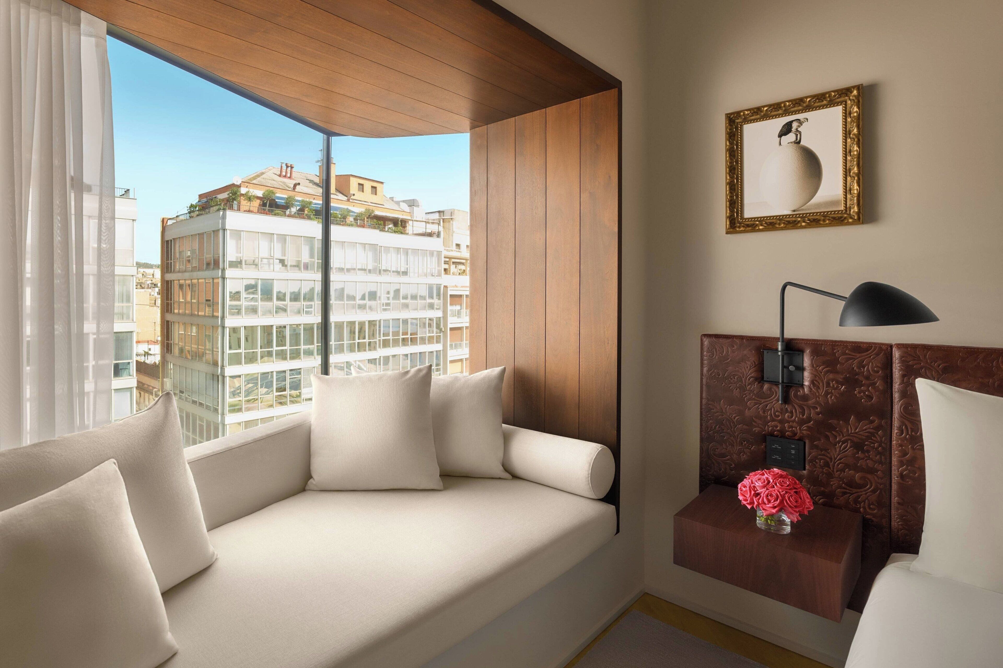 Una habitación de hotel moderna con un ventanal grande, un sofá con cojines, una mesita de noche con una lámpara y un cuadro enmarcado en la pared.