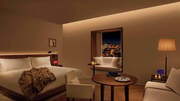 Habitación de hotel con cama, sofá, sillón, mesita y vista a un paisaje urbano nocturno.