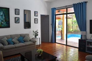 A living room with a sofa, a coffee table, and a TV stand. There is a door leading to a balcony with a pool. - 4 Bed Villa Private Pool BBQ Jomtien Beach Pattaya (Pattaya)