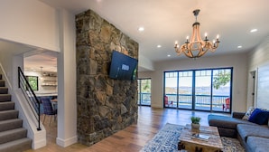 A spacious living area with a stone accent wall, a flat-screen TV, a chandelier, and a view of the outdoors.