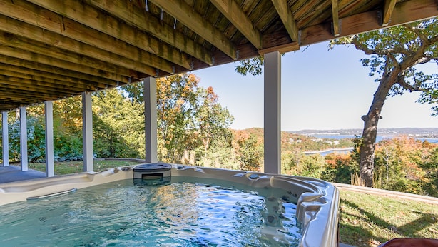 A hot tub under a wooden pergola with a view of a lake and autumn trees.