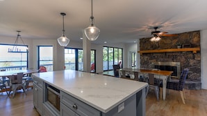 A spacious kitchen with a large island, pendant lights, a stone fireplace, and a view through large windows.