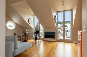Un salon moderne avec un canapé, une table ronde surmontée d’une lampe, un téléviseur et de grandes fenêtres donnant sur des immeubles.