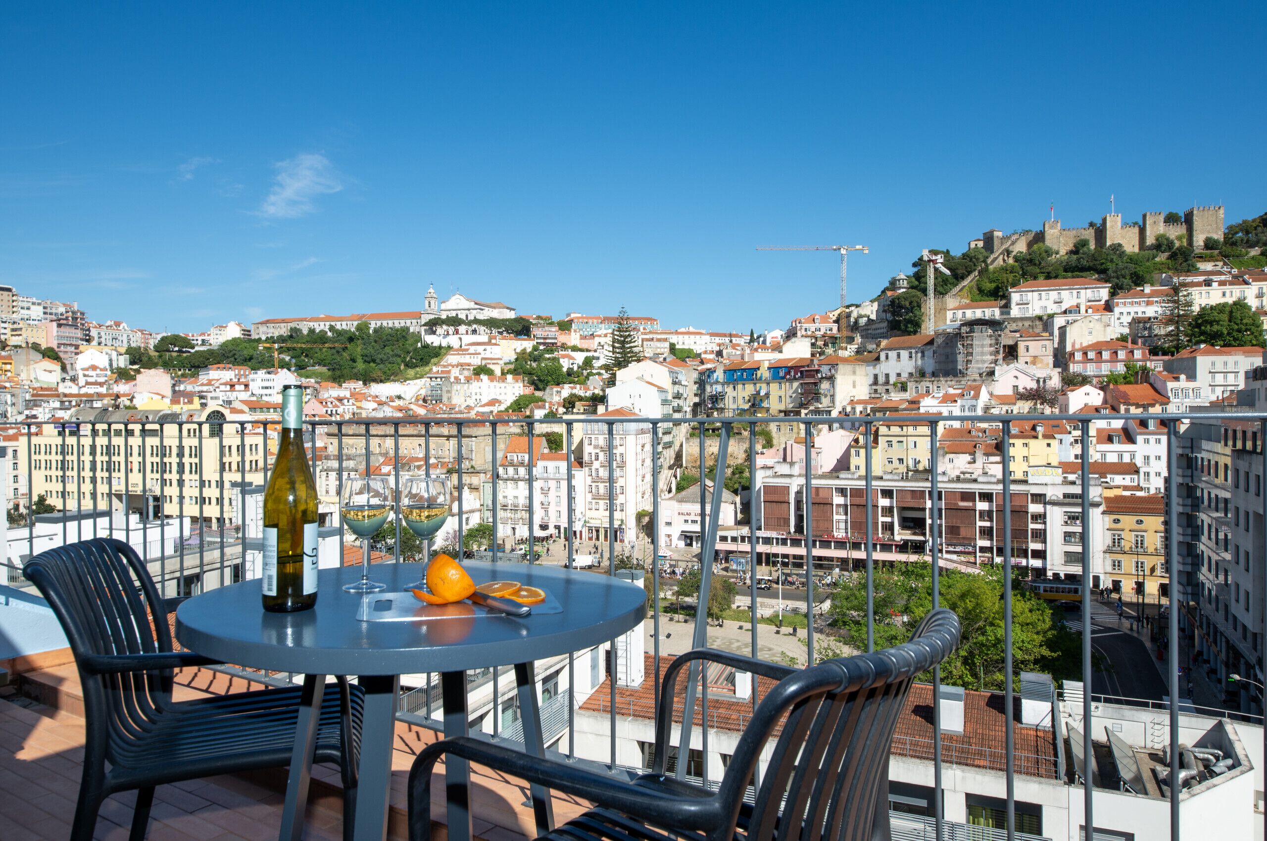Un balcón con una mesa, sillas, una botella y vasos, con vistas a un paisaje urbano que incluye edificios y un castillo.