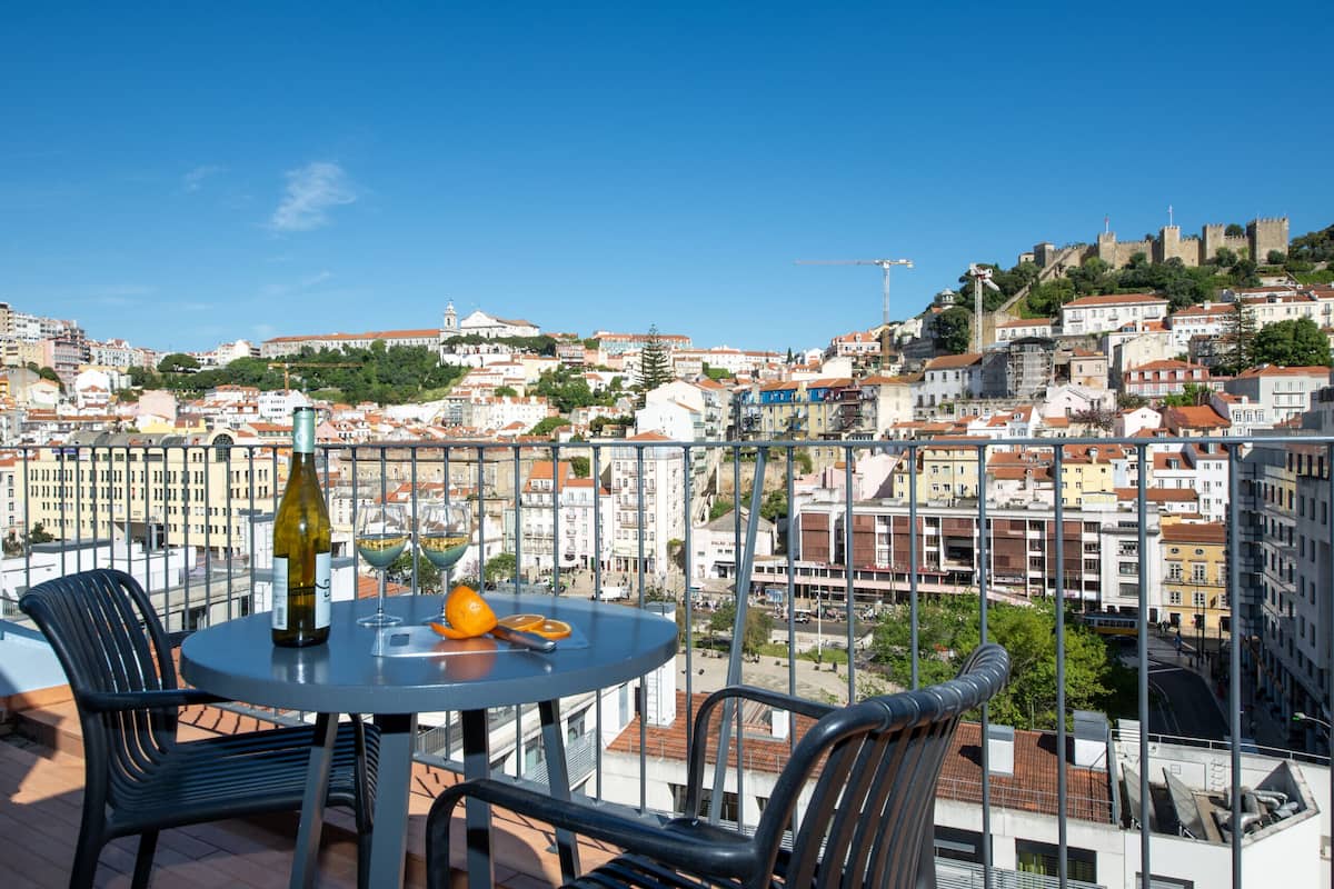 Un balcon avec une table, des chaises, une bouteille et des verres, donnant sur un paysage urbain avec des immeubles et un château.
