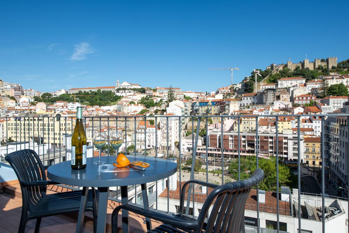 Un balcón con una mesa, sillas, una botella y vasos, con vistas a un paisaje urbano que incluye edificios y un castillo.