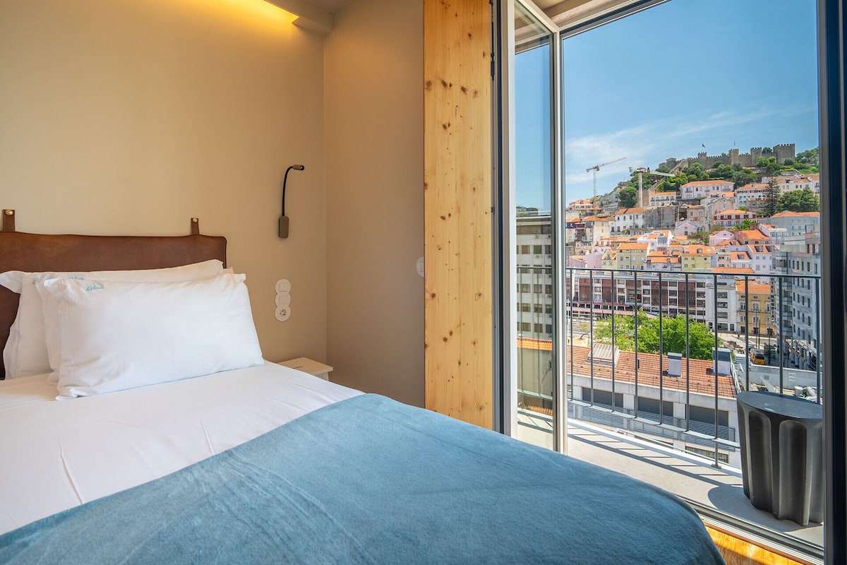 Hotelkamer met een bed, houten hoofdbord, nachtkastje, stopcontact en een groot raam met uitzicht op de stad.
