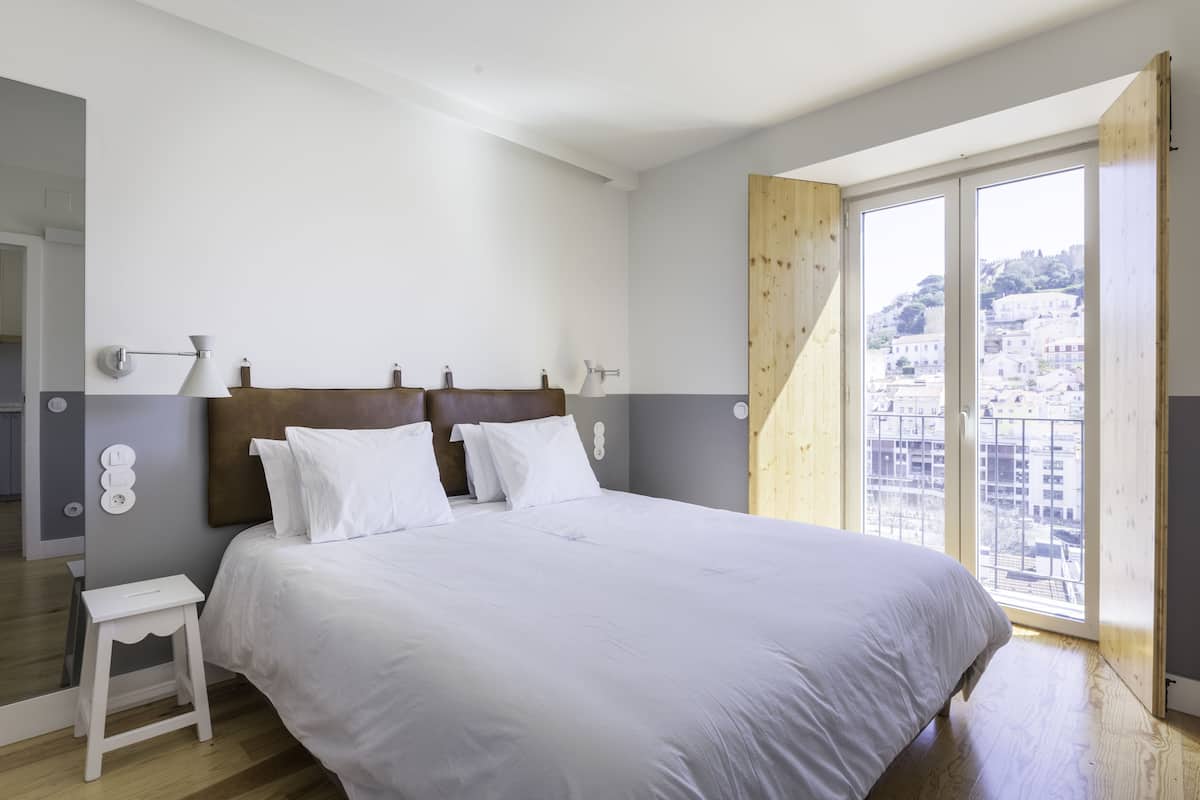 Une chambre avec un grand lit, une tête de lit en bois, une petite table de chevet blanche et une vue sur les immeubles par une grande fenêtre.