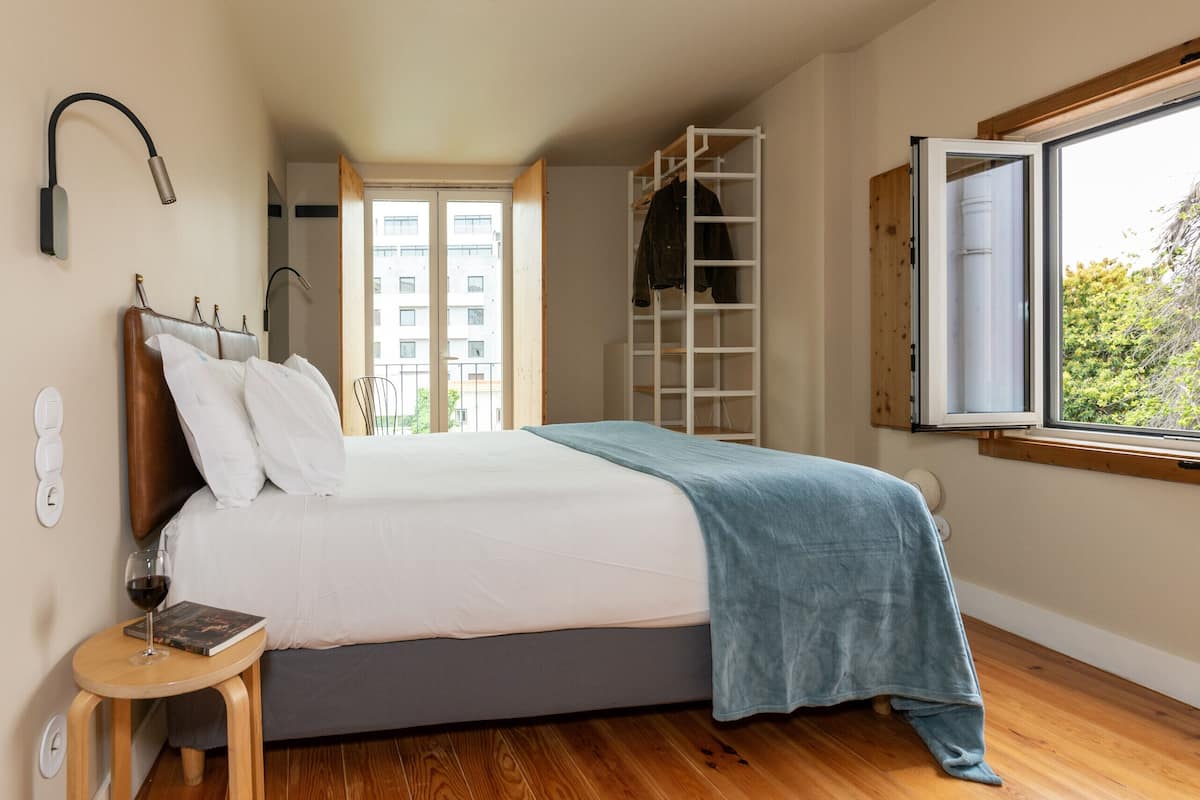 Une chambre à coucher avec un lit, une table de chevet sur laquelle se trouvent un verre à vin et un livre, une fenêtre donnant sur des immeubles et une échelle en bois.