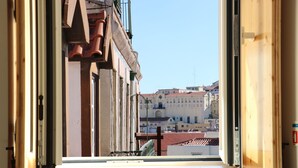 Blick durch ein offenes Fenster auf eine Stadtansicht mit Gebäuden und blauem Himmel.
