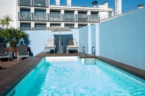 Ein Schwimmbecken mit Liegestühlen und Sonnenschirm, umgeben von einer blauen Mauer und einem Gebäude mit Balkonen.