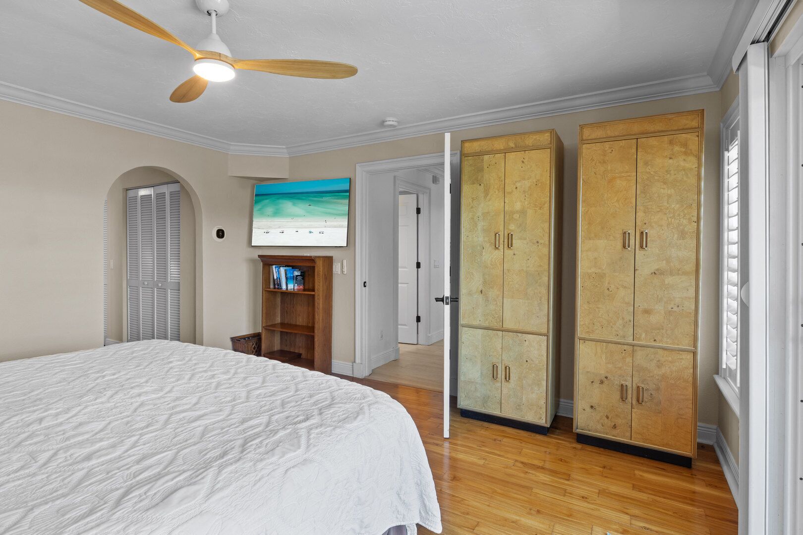 Une chambre à coucher comprenant un lit, une armoire en bois, un ventilateur de plafond, une bibliothèque et un tableau accroché au mur.