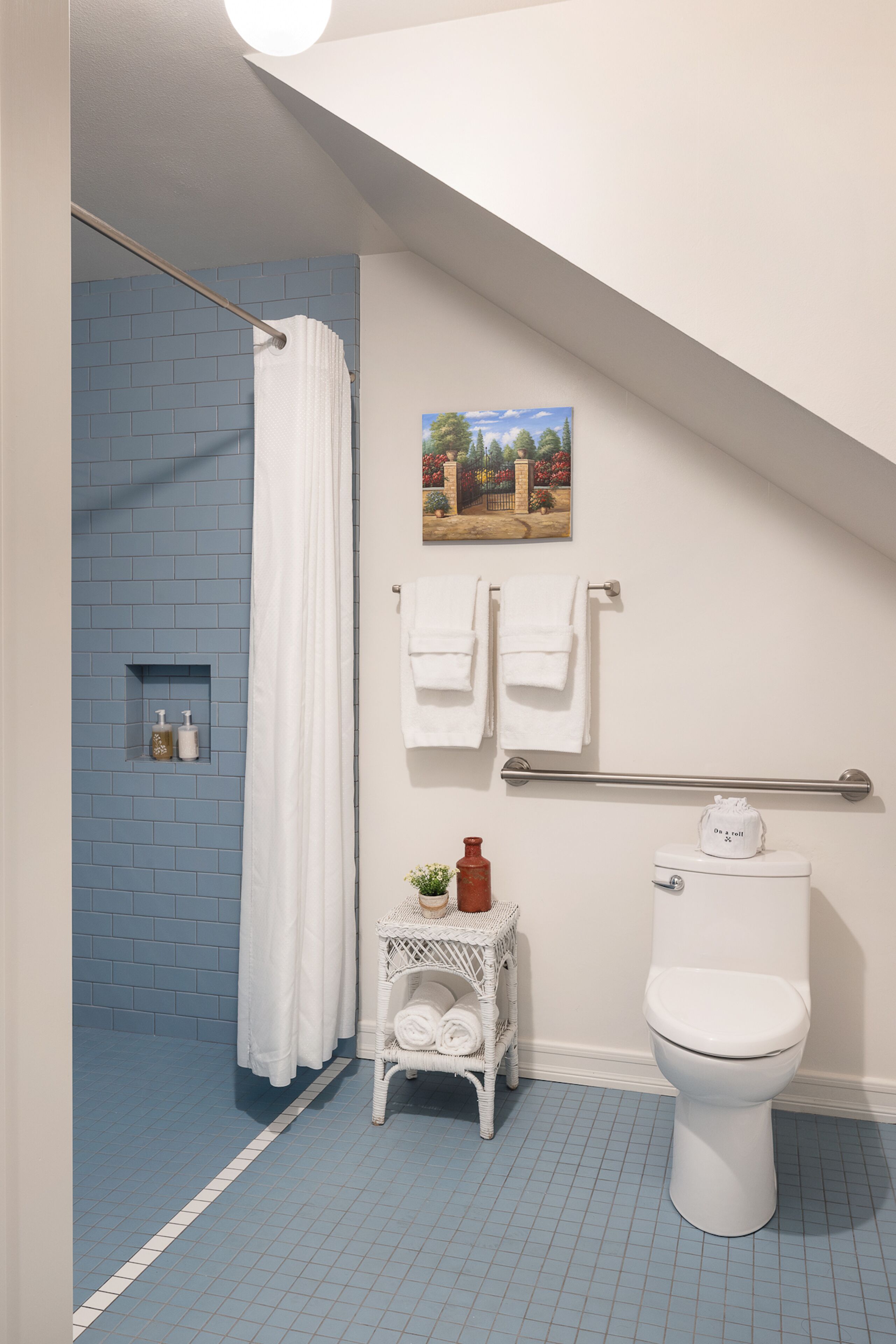 A bathroom with a blue-tiled shower, white fixtures, and a small white wicker side table with toiletries.