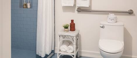 A bathroom with a blue-tiled shower, white fixtures, and a small white wicker side table with toiletries.