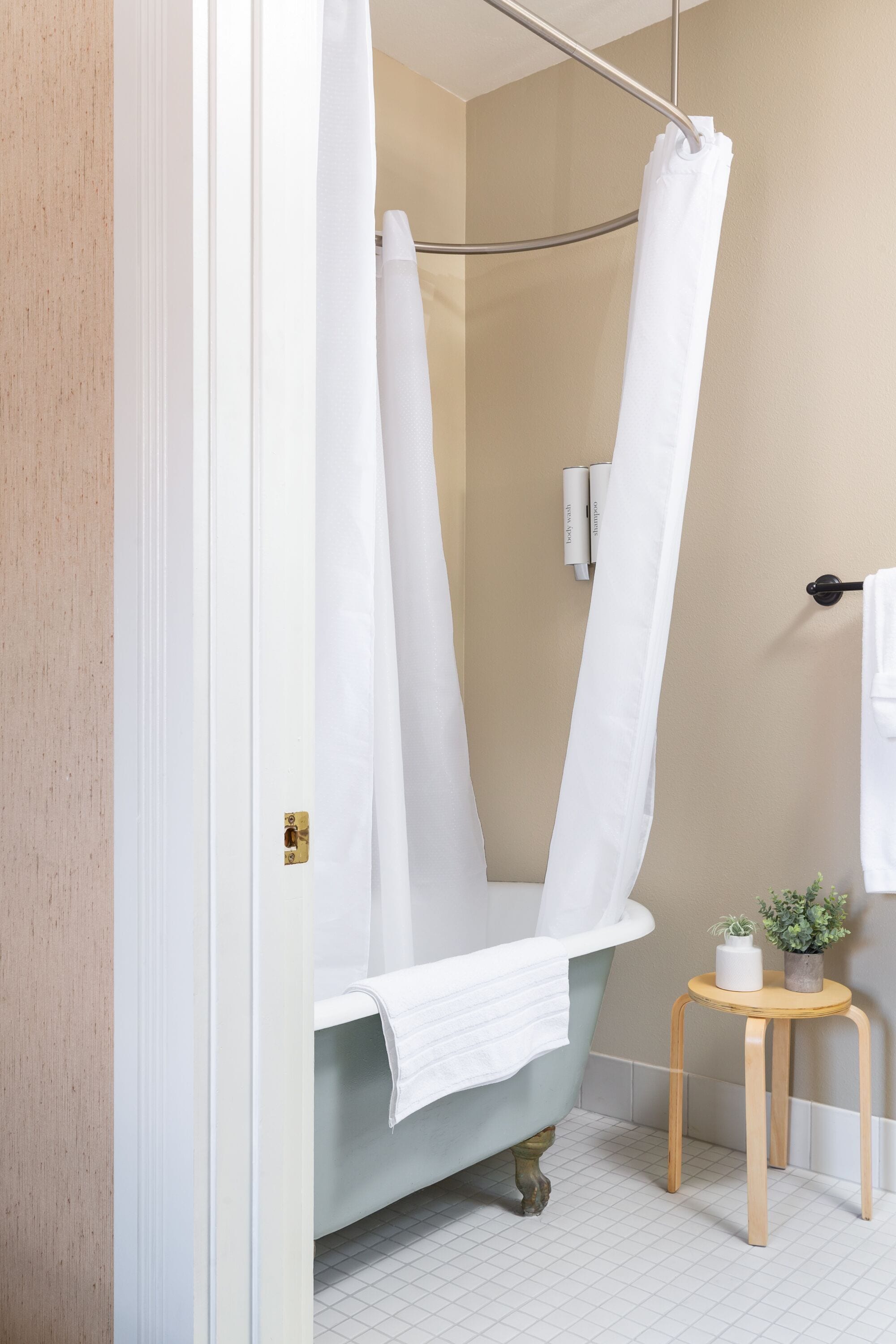 A bathroom with a clawfoot bathtub, a white shower curtain, a small wooden stool with a plant, and a towel rack.