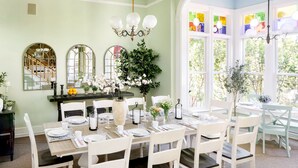 Comedor con una mesa grande dispuesta para seis personas, rodeado de sillas blancas, un candelabro y ventanas amplias con vidrieras.