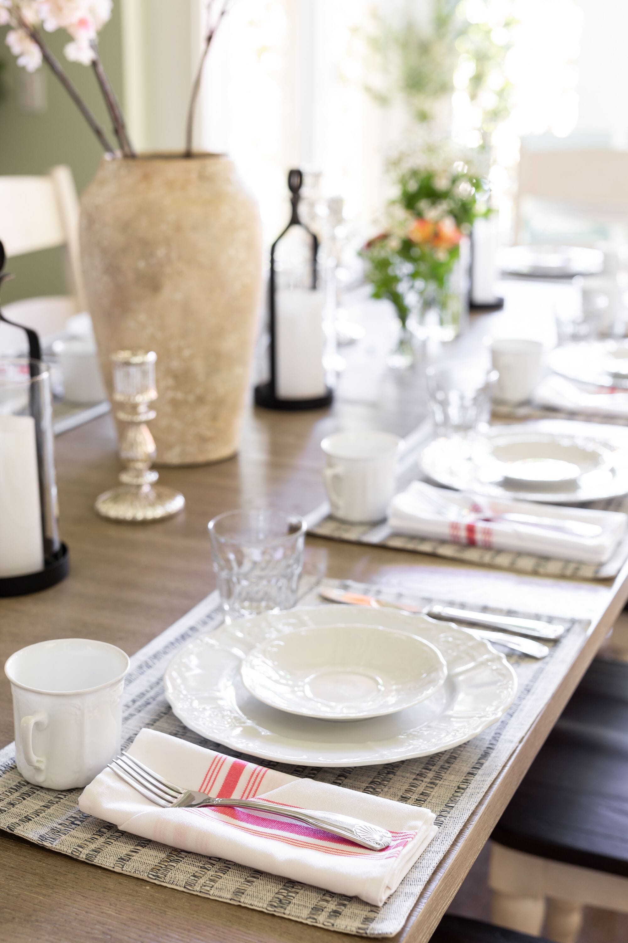 A neatly set dining table with white plates, cups, and cutlery, a vase with flowers, and a decorative candle holder.