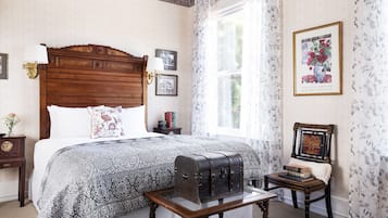 A bedroom with a wooden headboard, a patterned bedspread, a vintage suitcase, and a framed picture on the wall.