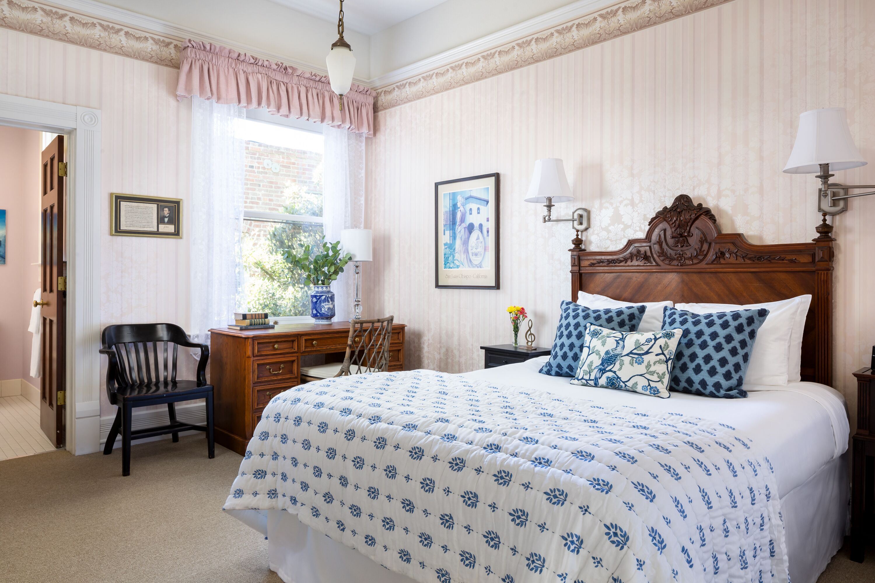 A bedroom with a large bed, a wooden desk, a chair, and a window with curtains.