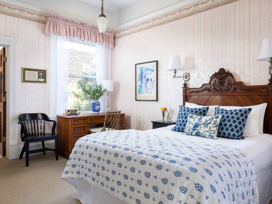 A bedroom with a large bed, a wooden desk, a chair, and a window with curtains.