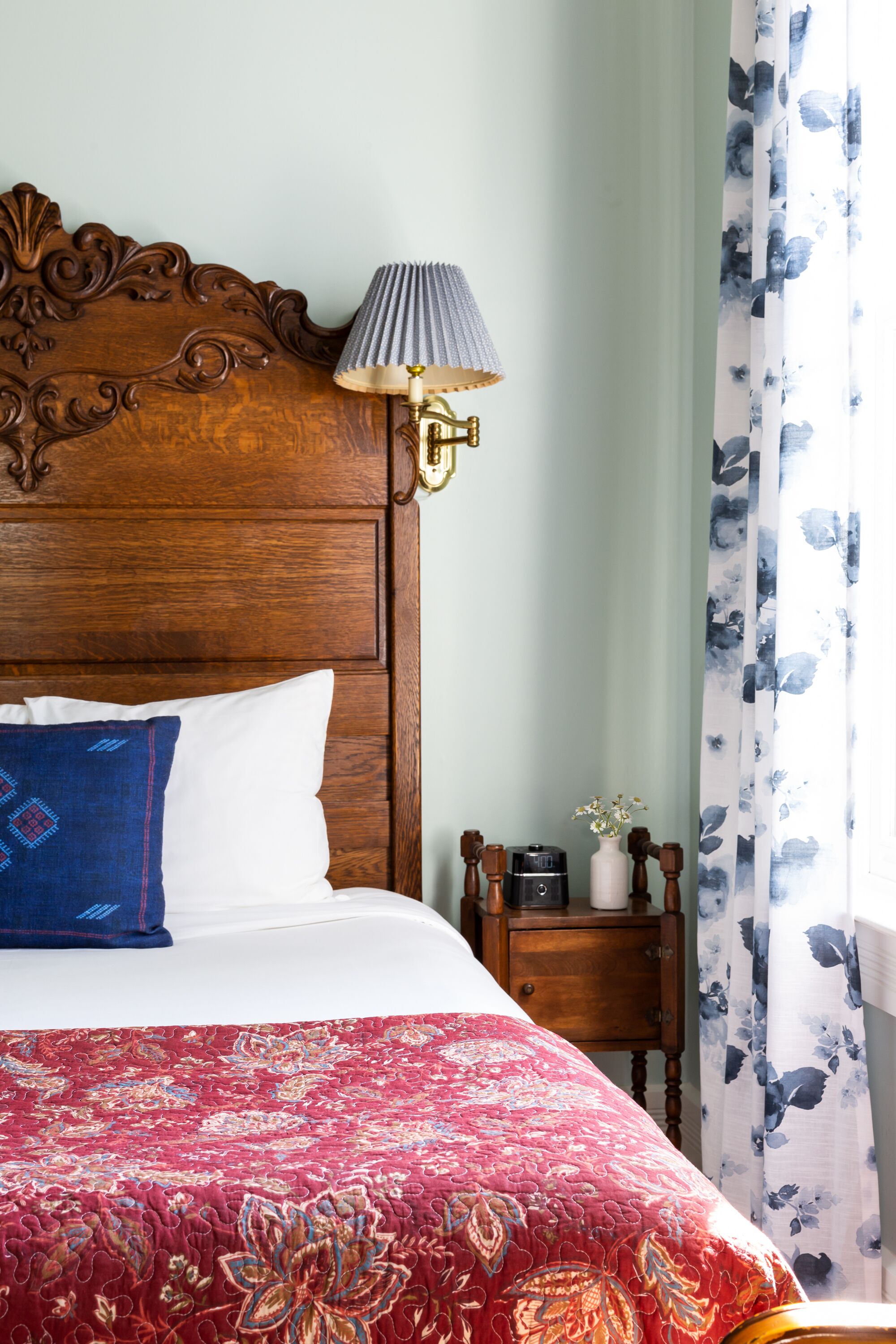 A bed with a red floral bedspread, a wooden headboard, a white pillow, a blue pillow, a bedside table with a lamp, and a floral curtain.