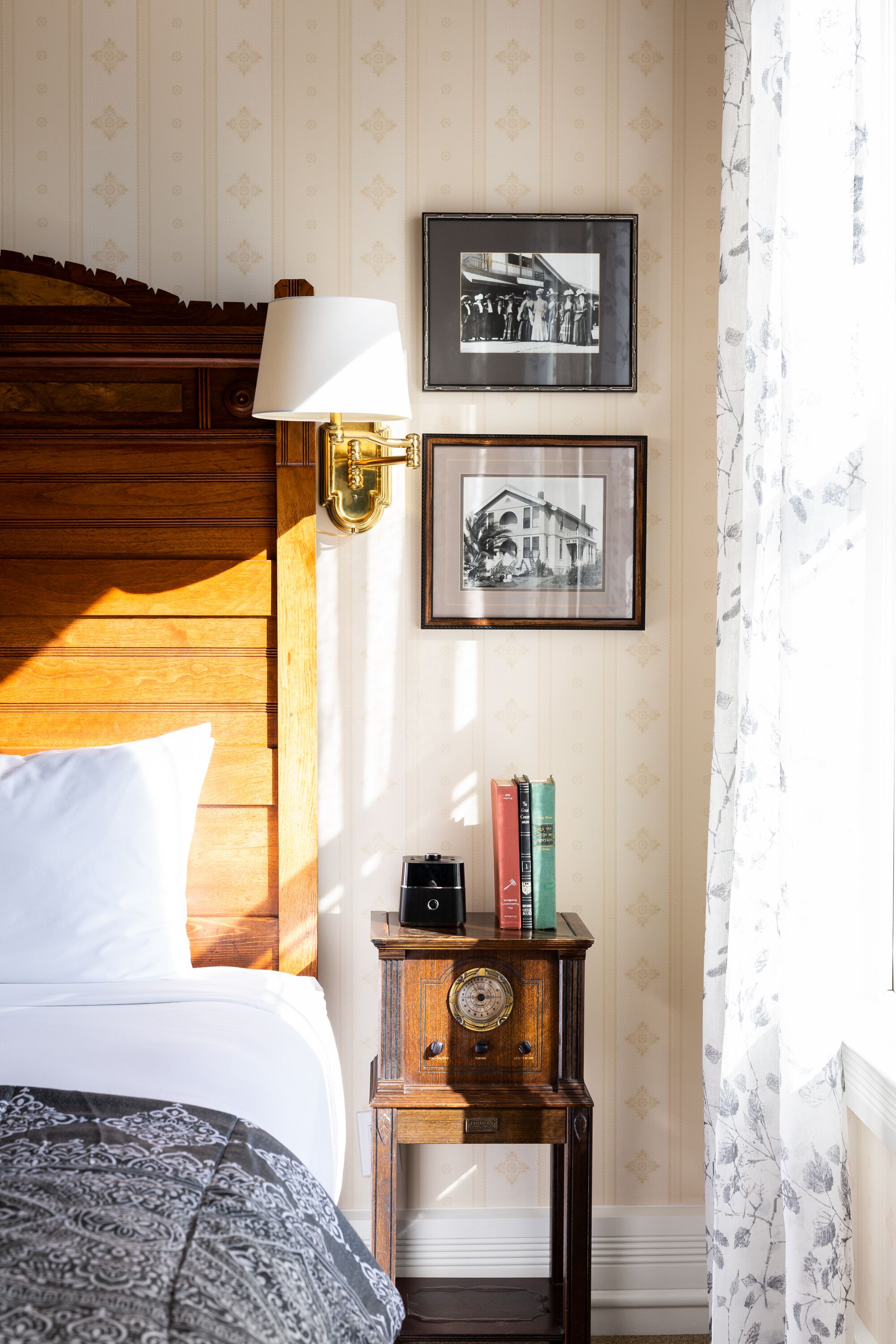 Ein ordentlich bezogenes Bett mit einer gemusterten Decke, ein Nachttisch mit einer Uhr und zwei gerahmte Fotografien an der Wand.