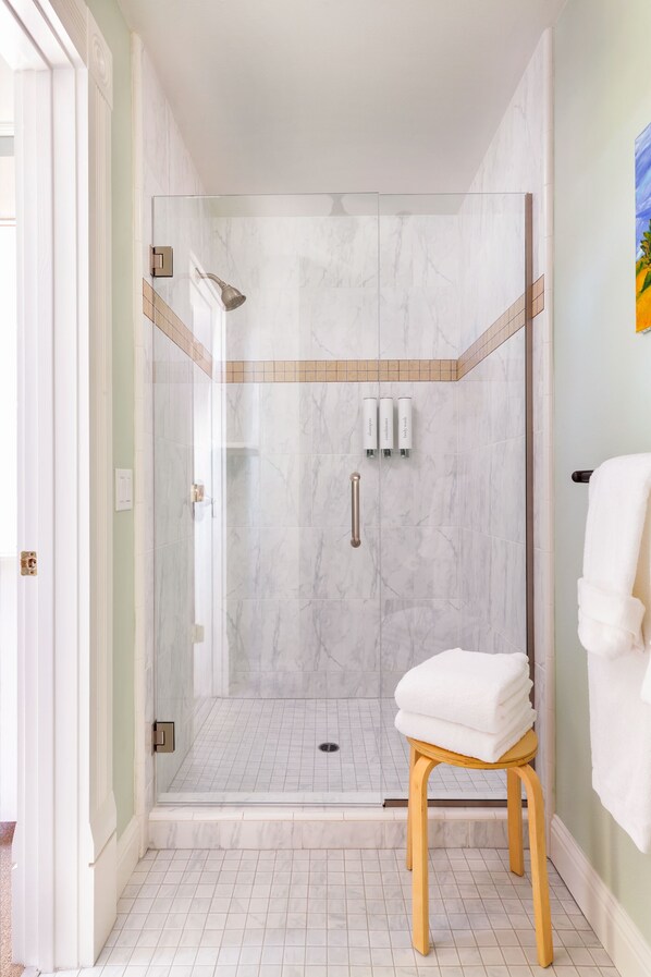A modern bathroom with a glass-enclosed shower, a wooden stool with a folded towel, and a tiled floor.