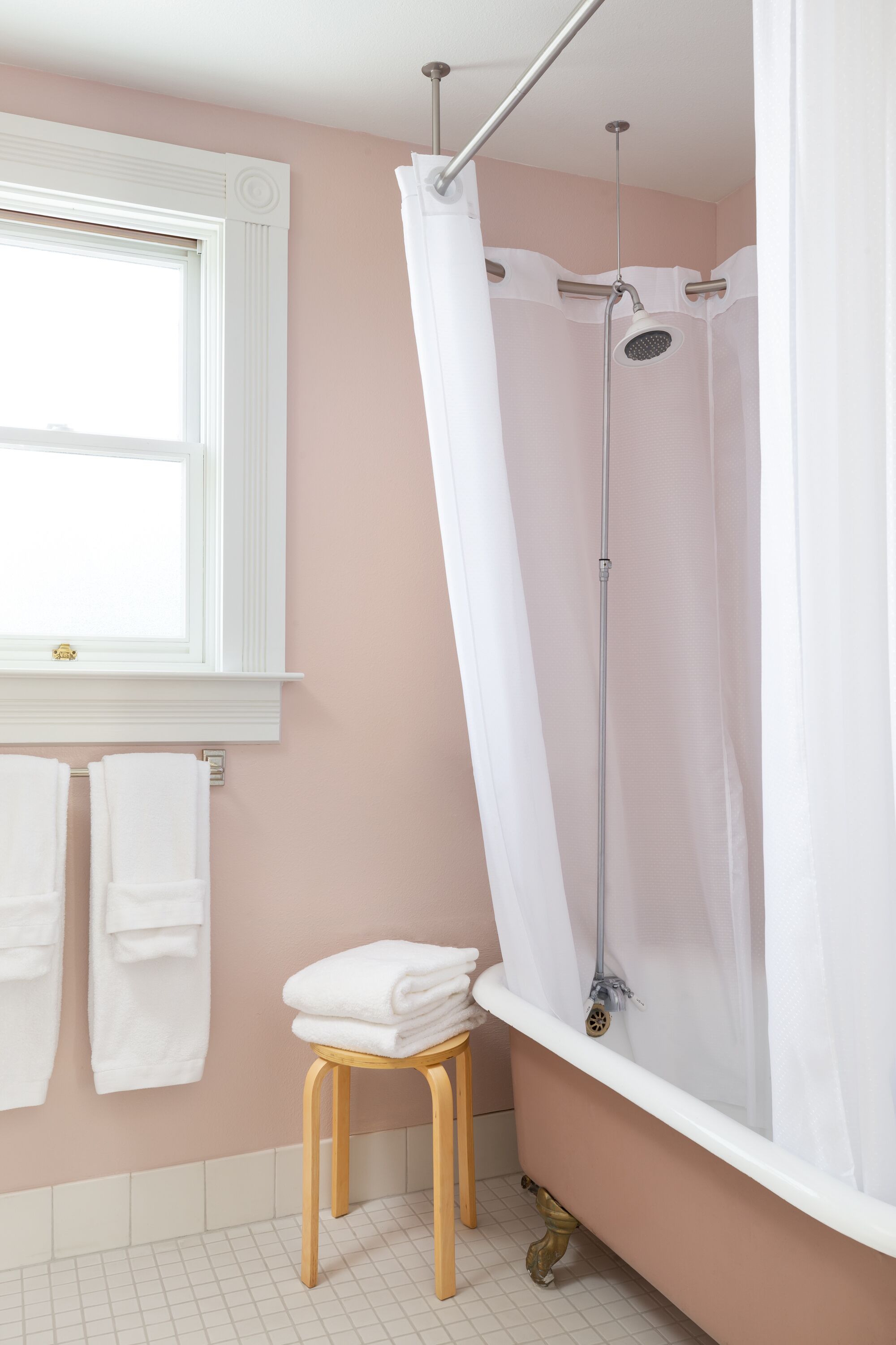 A bathroom with a bathtub, a shower curtain, a window, and a stool with towels.