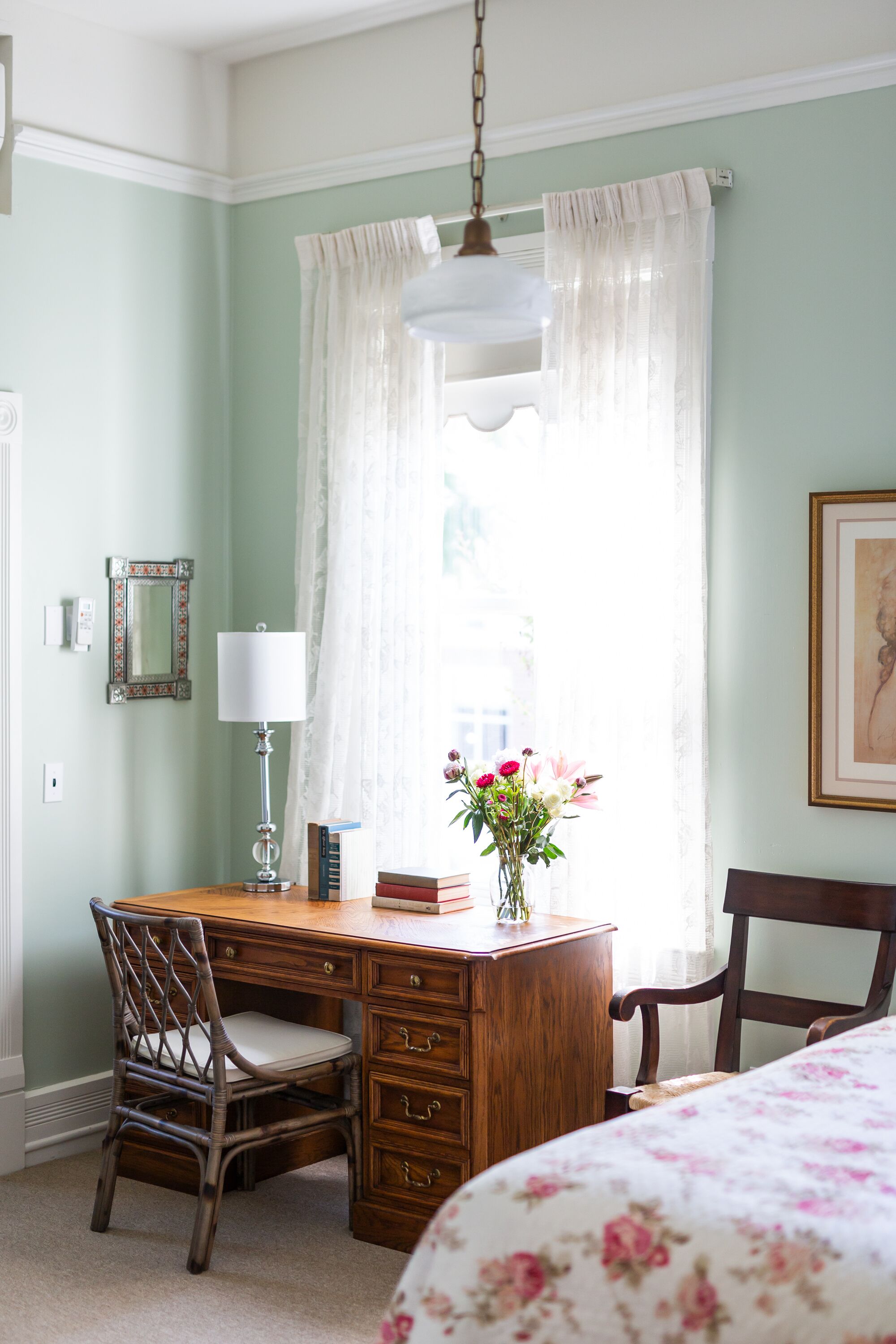 A room with a wooden desk, a chair, a lamp, a vase with flowers, and a bed with a floral bedspread.